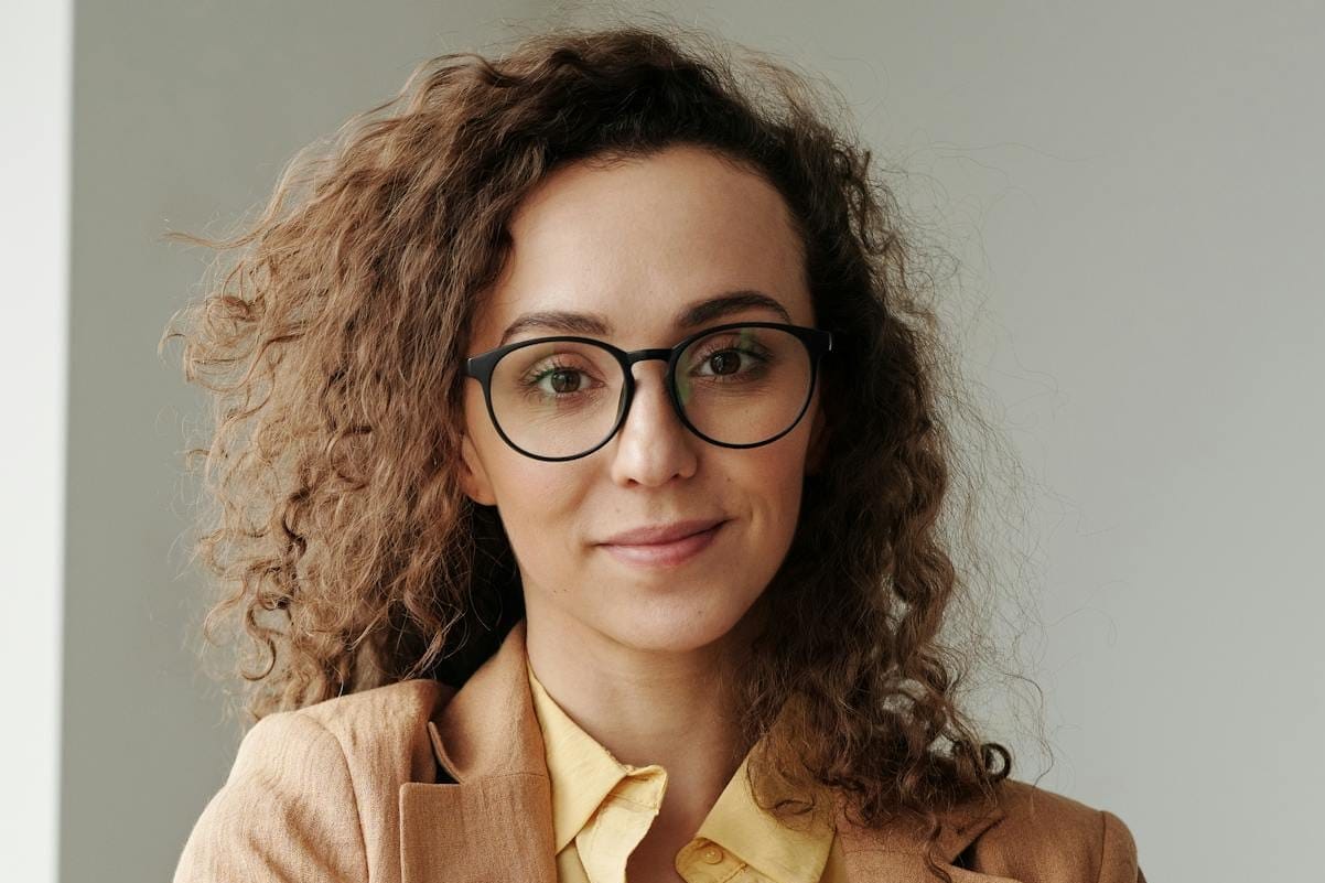 Curly-haired businesswoman wearing glasses holds a laptop in a bright office setting.